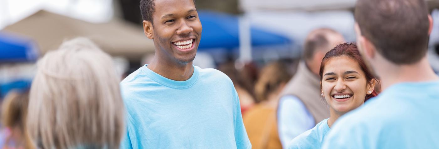 A group of young adults wearing matching light blue tee shirts smile and talk to one another at a community event.