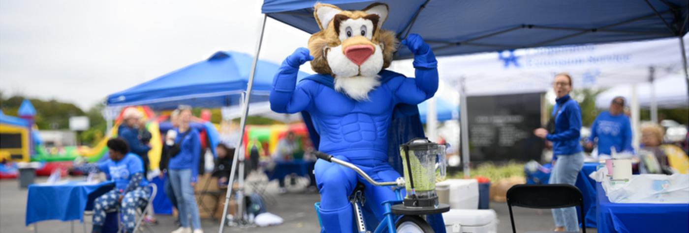 Wally Cat mascot flexes his biceps while sitting on a bicycle.