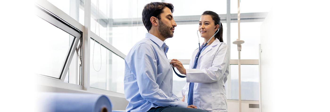 A femal doctor in a white coat uses a stethoscope to listen to the chest of her patient, an adult male in his 30s.