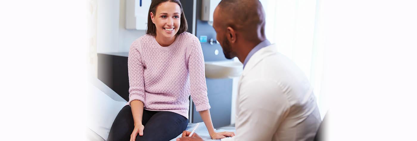 A woman wearing a pink sweater smiles as she speaks with her doctor, an African-American man wearing a white coat. 