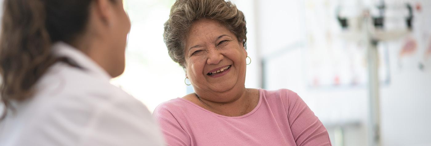 An older woman wearing a pink blouse smiles and she speaks with her provider, a woman (seen from behind), wearing a white coat. 