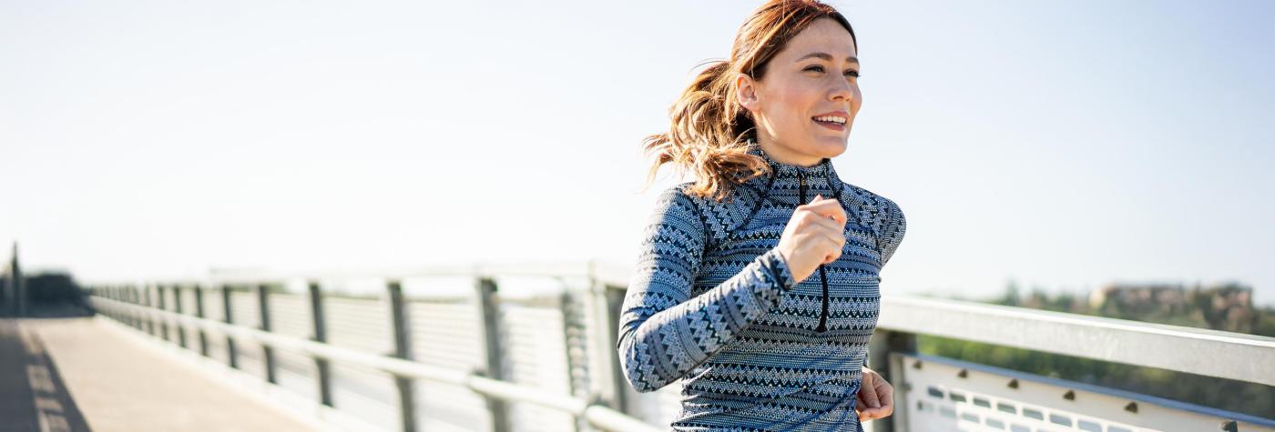 A woman wearing a blue striped jacket jogs along the sidewalk of a bridge.