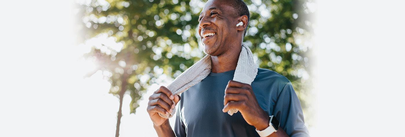 A smiling middle aged African American man wearing a blue tee shirt pauses during his outdoor workout. He has an earbud in and a white towel draped over his shoulders. 