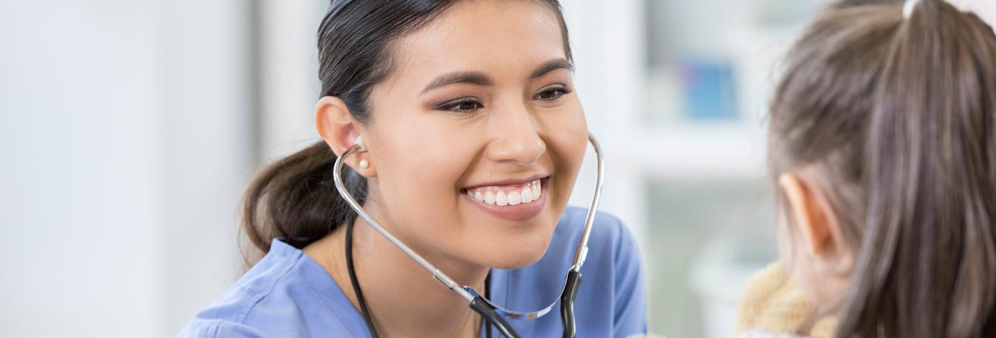 A female provider wearing a light blue scrub top and stethoscope smiles as she interacts with a young female patient.