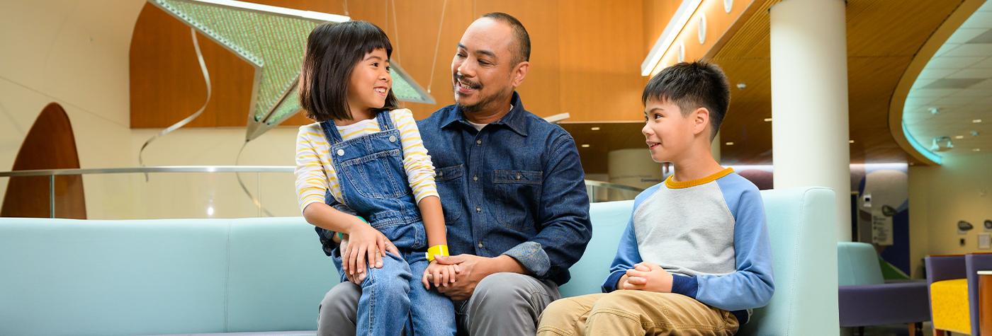 A brother and sister sit together with their father in a waiting area at Kentucky Children's Hospital.