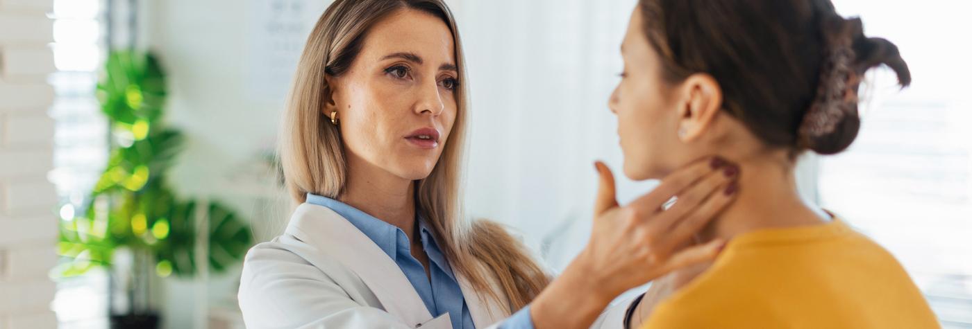 A female doctor wearing a white coat palpates the lymph nodes on the side of a young woman's neck.
