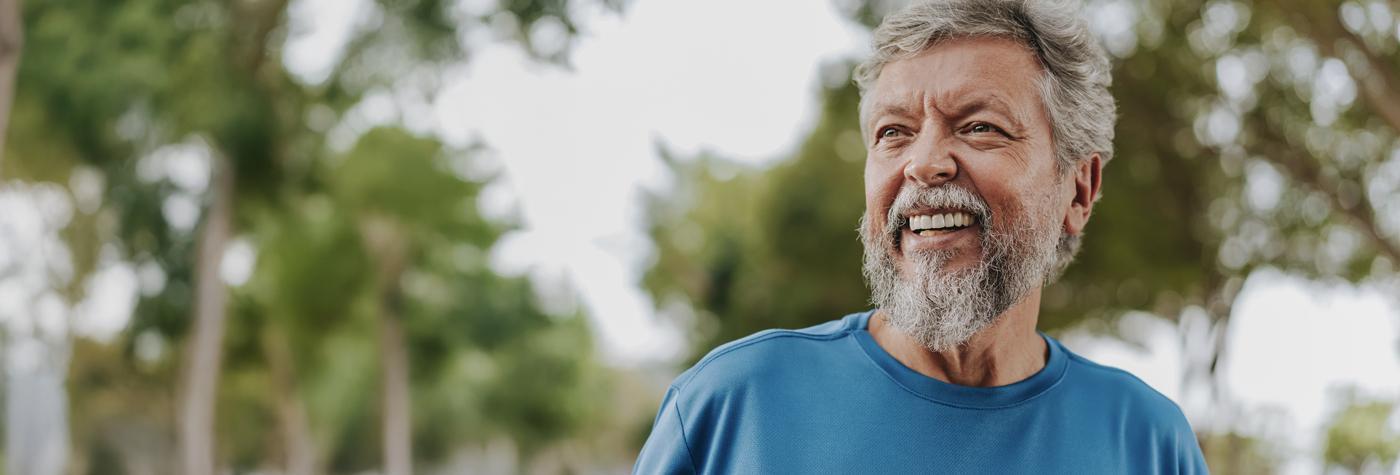 A man with gray hair and a gray beard smiles with trees in the background. He is wearing a teal blue tee shirt.
