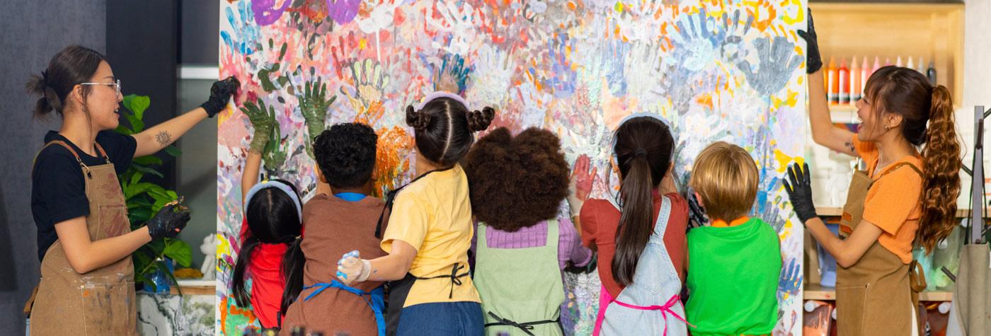 A group of children, assisted by two adults in light-brown aprons, paint a large canvas together.