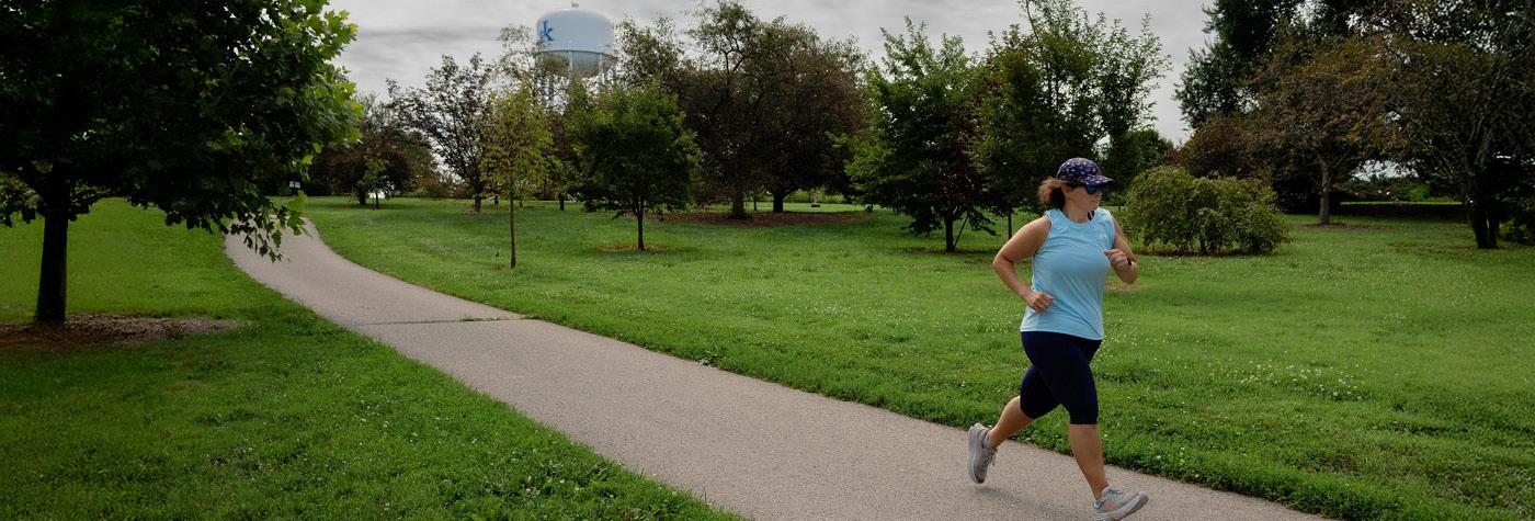 A woman wearing a baseball cap and light blue shirt jogs along a path in the UK Arboretum.