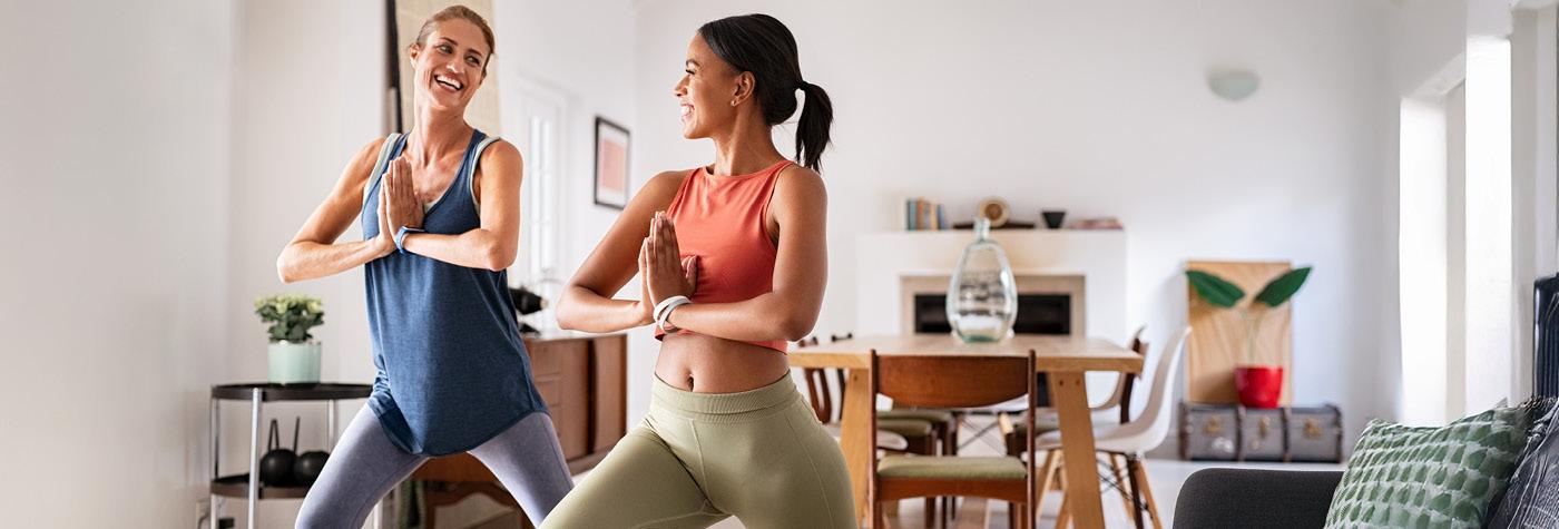Two women wearing workout clothes smile as they stand side by side doing yoga poses. 