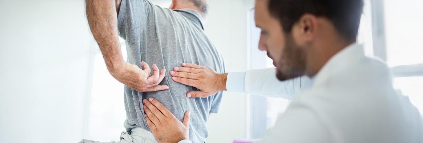 A male doctor wearing a white coat examines a male patient's back. The patient is wearing a light grey polo shirt. 