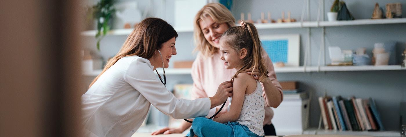 A female doctor wearing a white coat uses a stethoscope to listen to a young girl's heart while her mother looks on, smiling.
