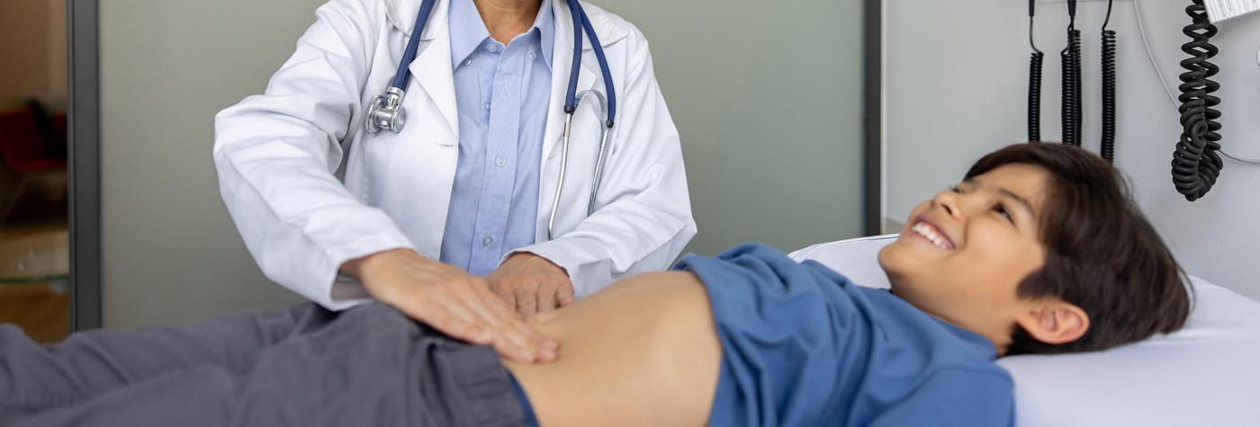A boy wearing a blue shirt lies on an exam table while a doctor, dressed in a white coat with a stethoscope around their neck, palpates the boy's abdomen.