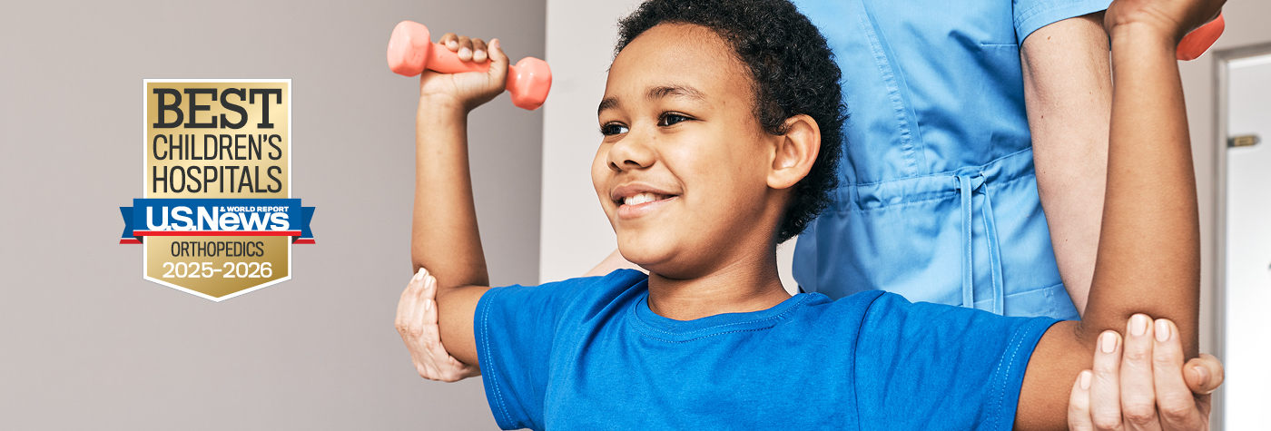 A boy in a blue shirt flexes his biceps and holds small hand weights while a person in blue scrubs stands behind him, supporting his elbows. A badge superimposed on the image reads "US News and World Report, Best Children's Hospitals, Orthopedics, 2025-2026."
