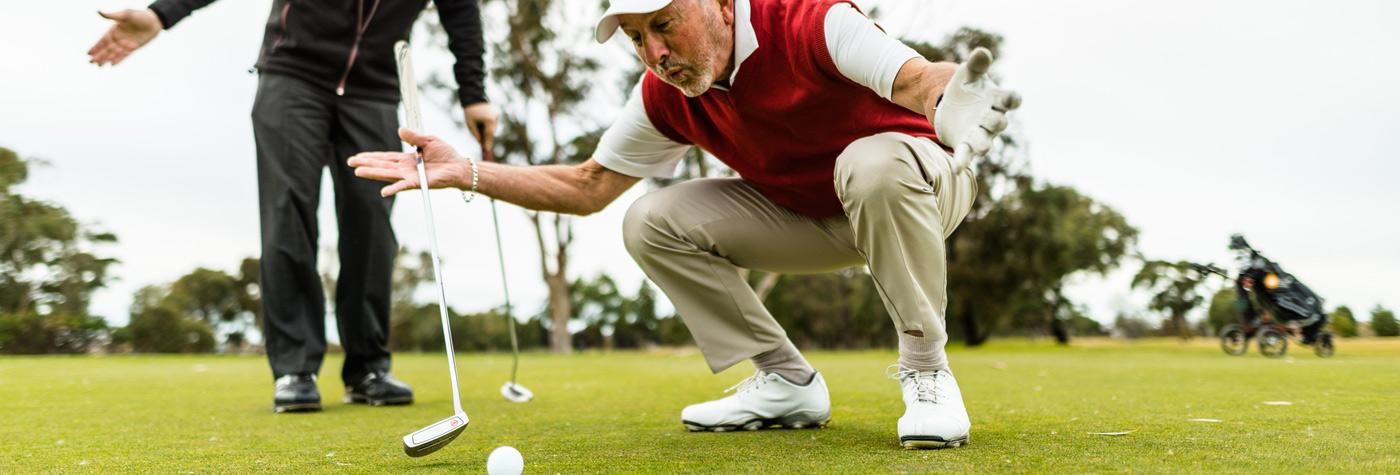 An older man wearing a red vest squats down next to a golf ball on a golf course.