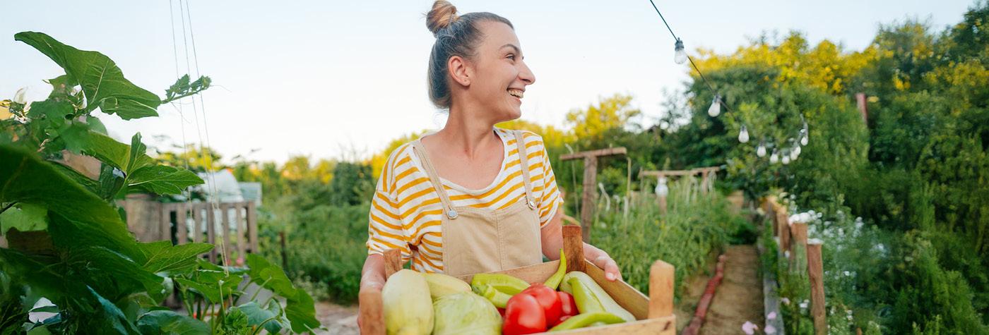 A woman wearing a yellow and white striped shirt and overalls carries a basket of vegetables through a garden.