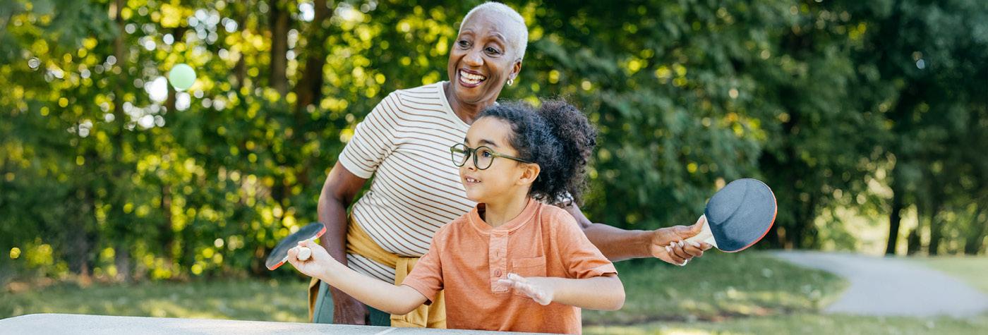 An older woman plays ping pong alongside a young girl. Both are smiling.