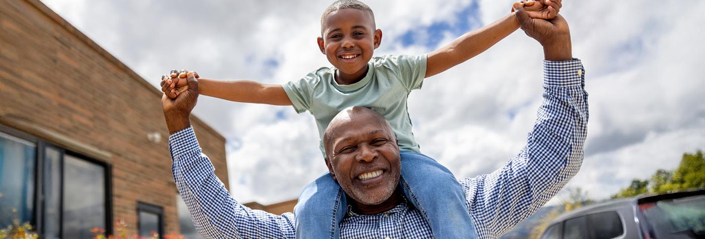An older man carries a young boy on his shoulders. They are both smiling.