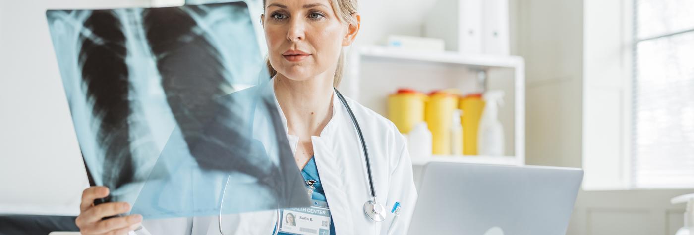 A female doctor, wearing a white coat with a stethoscope draped around her neck, reviews an X-ray while seated at her desk. 
