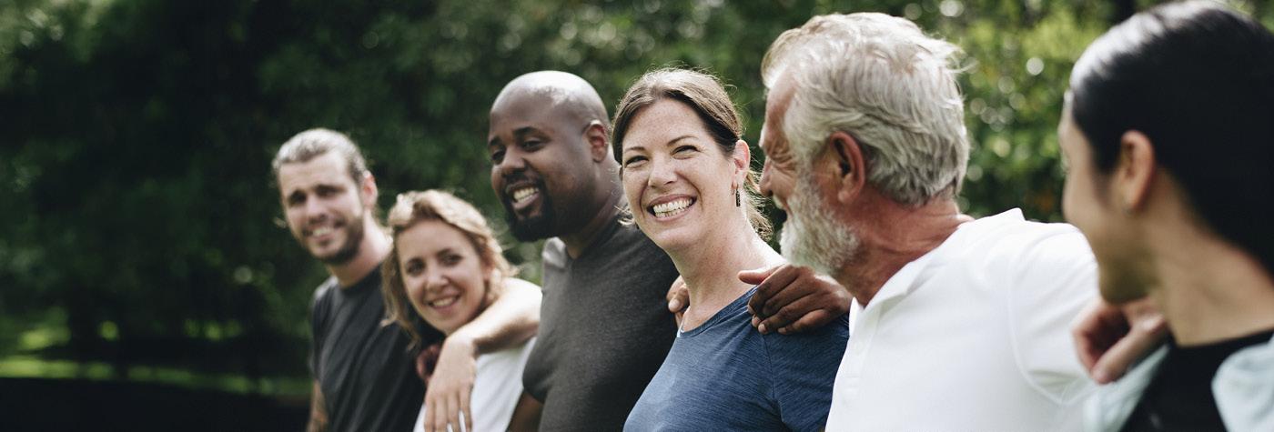 A diverse group of adults stands side by side with their arms over each others' shoulders.