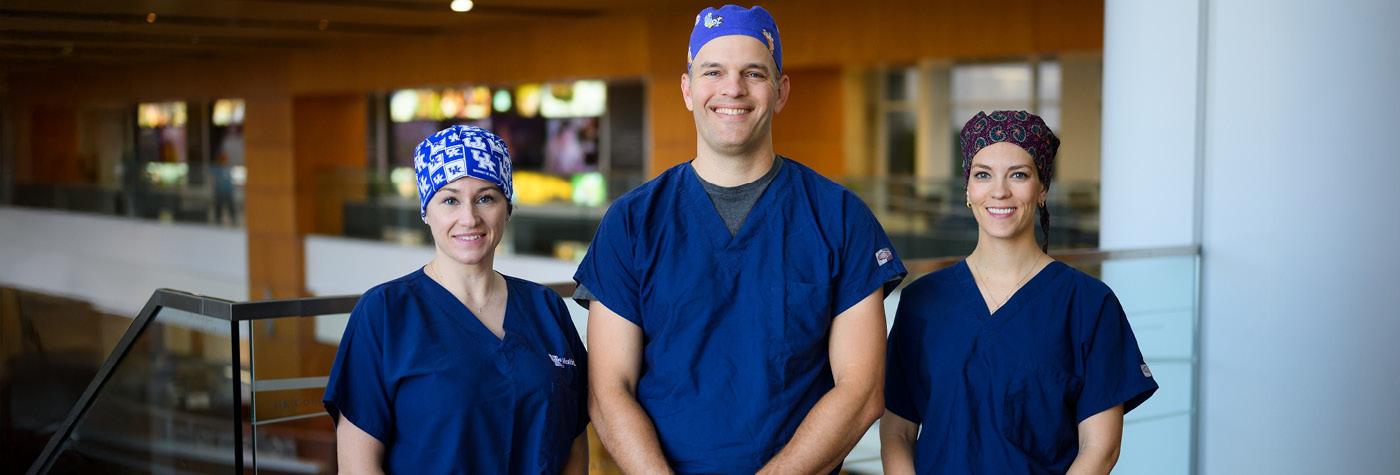 Three doctors wearing scrubs and scrub caps stand side by side smiling.