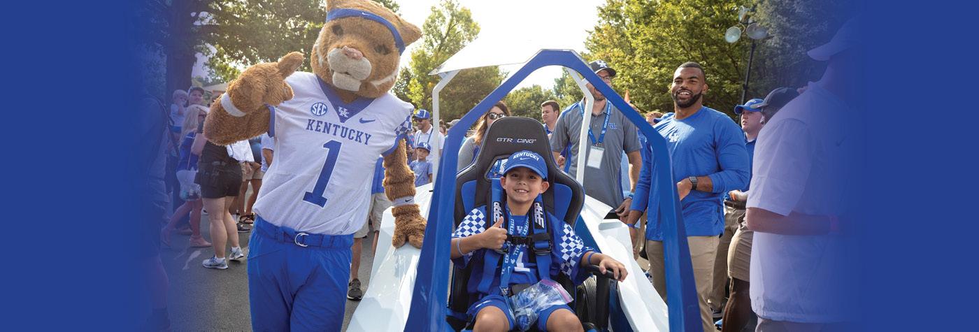 A young boy rides in the Lift Them Up cart, accompanied by the UK wildcat mascot.