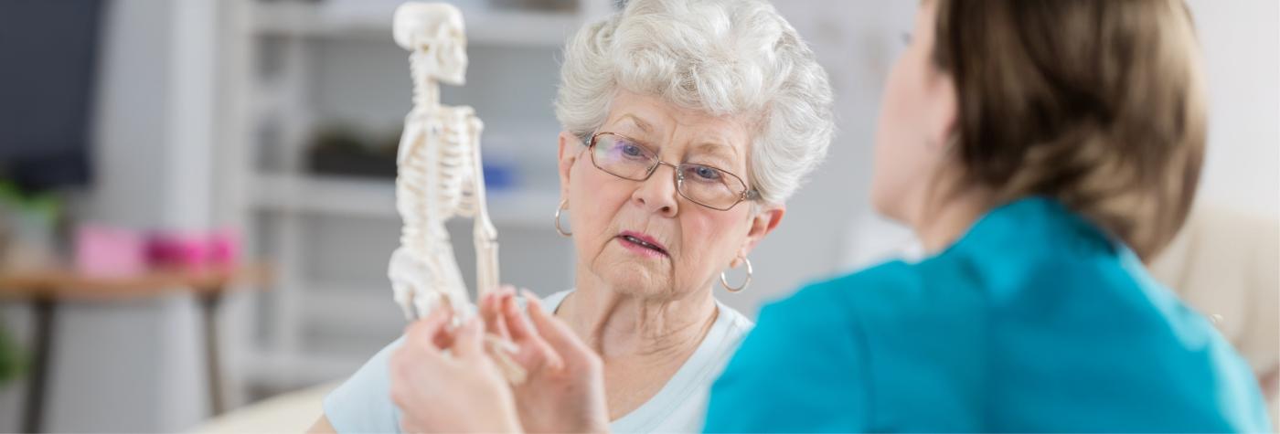 A doctor wearing teal scrubs shows an older woman a small figurine of a human skeleton.