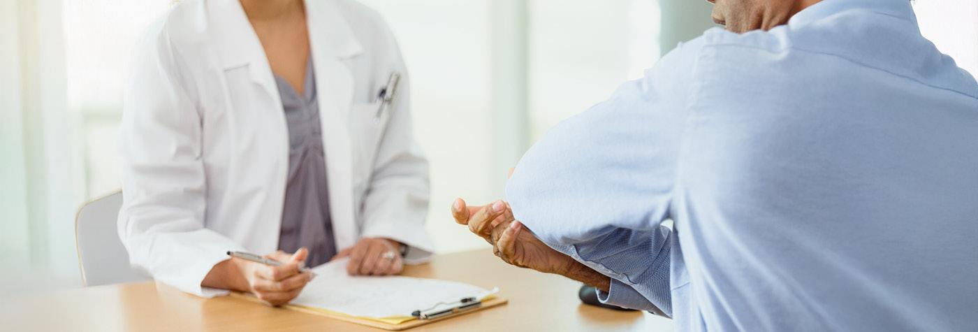 A doctor wearing a white lab coat speaks with a patient who is wearing a blue shirt and pointing to his bent elbow. We only see the subjects of the photo from the neck down to just below the chest.