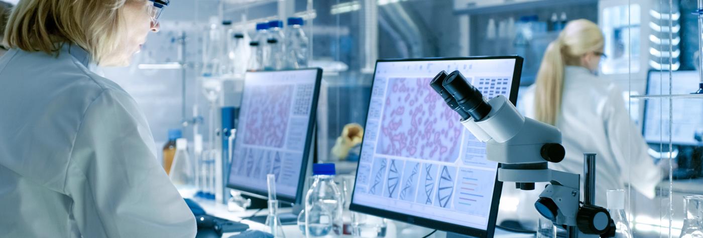 A researcher works at a laboratory bench with microscopes and computer screens displaying cellular and genetic imagery.