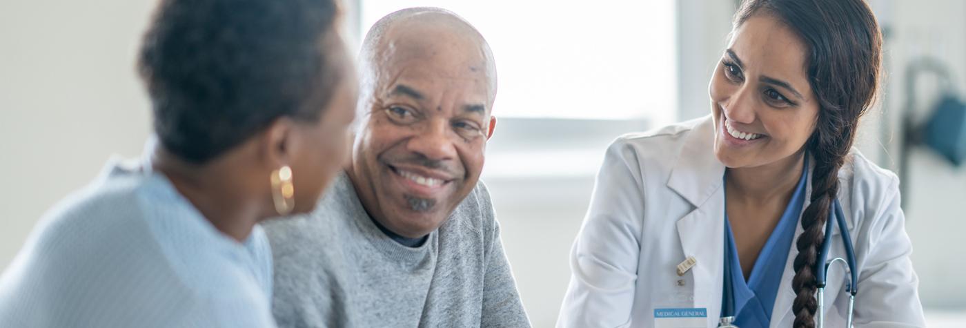 Healthcare provider meets with two adults during a clinical consultation in a medical exam room.