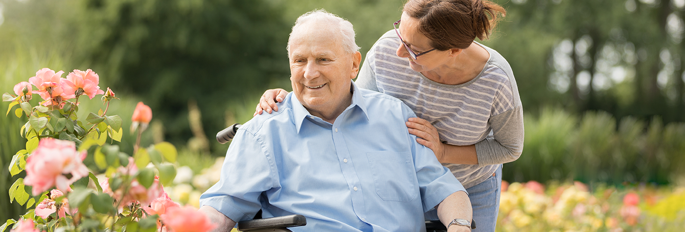 A person seated in a wheelchair outdoors among flowering plants, with another person standing behind the wheelchair and resting a hand on the seated person’s shoulder. Pink roses and greenery are visible in the foreground and background.