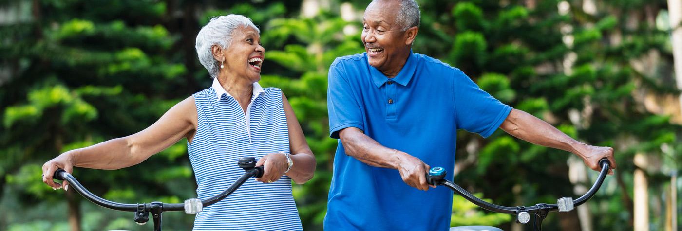 Two adults stand side by side outdoors, each holding the handlebars of a bicycle, with green trees in the background, suggesting active movement and wellness in a natural setting.