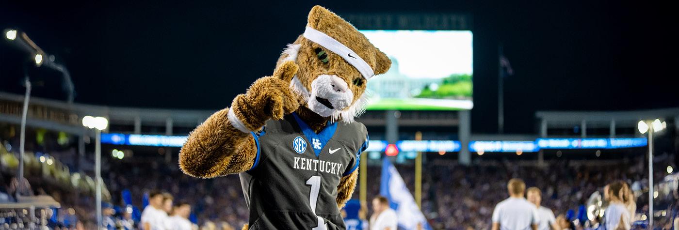 The UK Wildcat mascot poses for a photo holding up a number-one sign with his finger during a night game at Kroger Field. Members of the UK cheerleading squad are visible in the background.