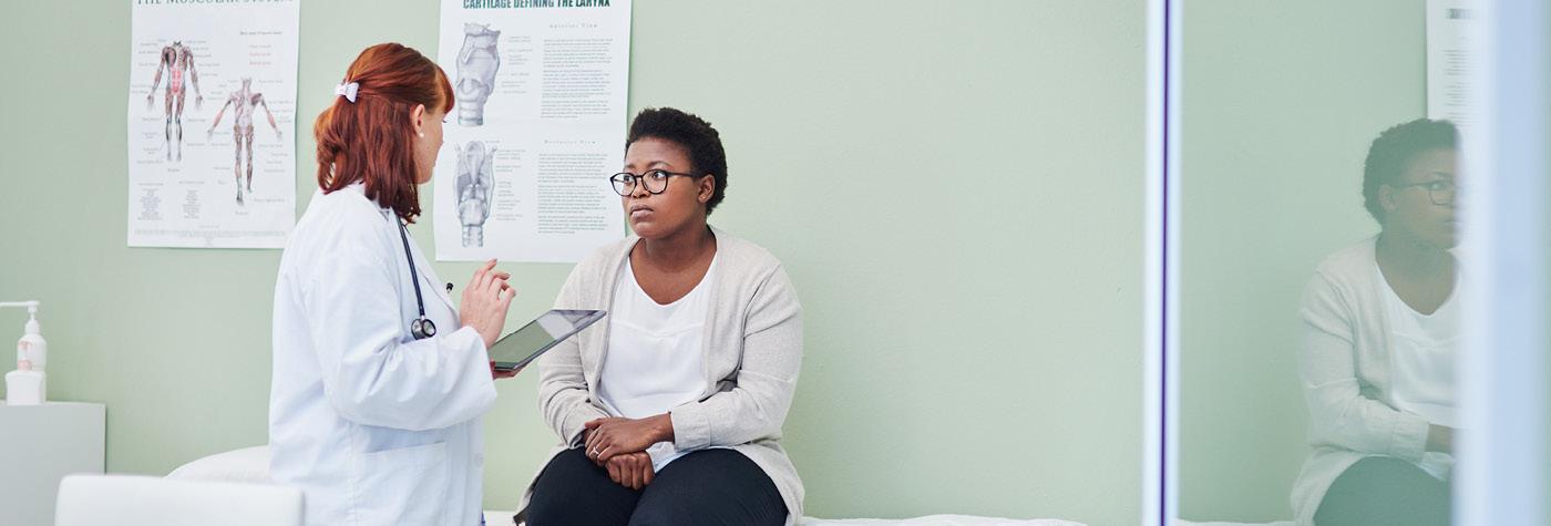Healthcare provider speaks with a seated patient in an exam room, referencing a tablet, with anatomical charts visible on the wall behind them.