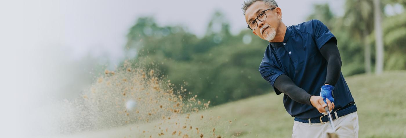 A man in a blue shirt hits a golf ball out of a sand trap, causing sand to fly everywhere in the foreground of the shot.