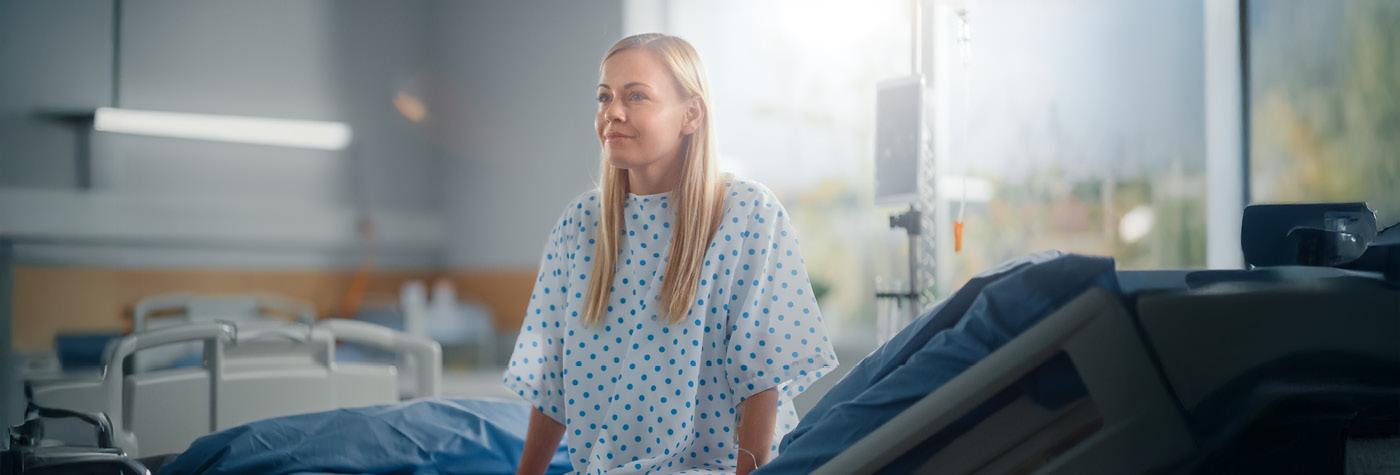 A woman wearing a hospital gown sits on the edge of a hospital bed.