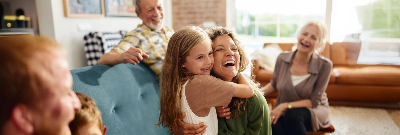 A group of extended family members smile and laugh as they spend time together in a well-lit living room with many windows.