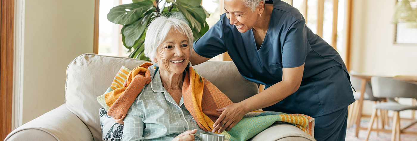A caregiver wearing scrubs drapes a blanket around the shoulders of a woman sitting in an armchair.