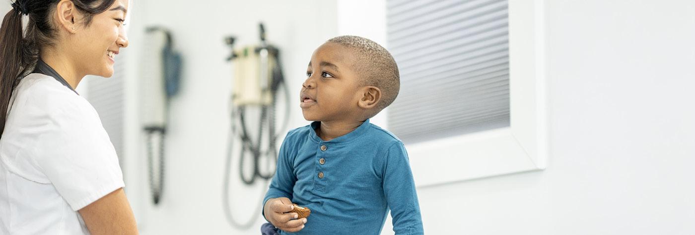 A female medical provider wearing a white scrub top and a stethoscope around her neck smiles at a young boy wearing a blue shirt seated on an exam table.