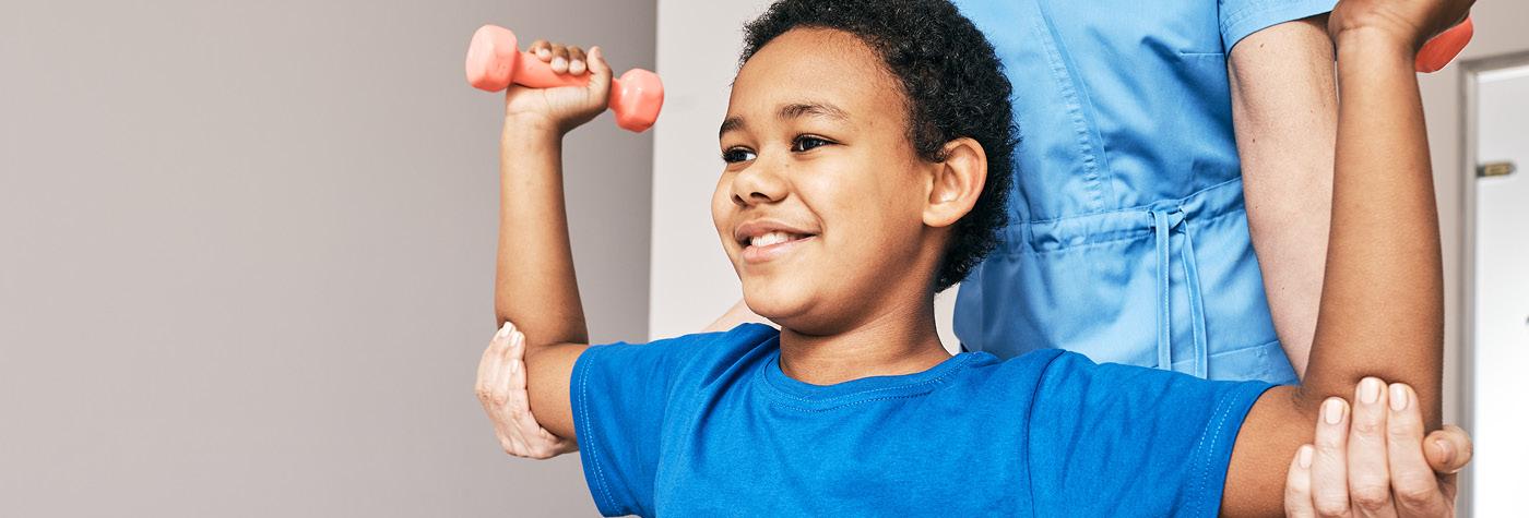 A boy wearing a blue tee shirt lifts two small hand weights while being assisted by a person wearing a light blue scrub top.