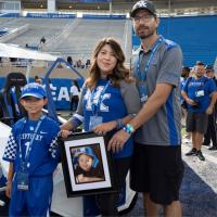 Liz, Benjamin, and Maximo prepare to lead the UK Football season opener, the Cat Walk. Liz holds a framed portrait of Marco.
