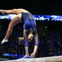 Mac flips backwards atop a balance beam, wearing a sparkly black and blue leotard and a blue bow in her hair.