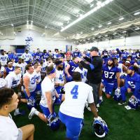 Coach Stoops talks to his team inside the UK football practice facility. He is surrounded by players, who are kneeling down around him.