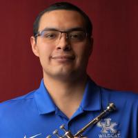 Adiel Nájera holds his trumpet while standing in front of a red backdrop. He is wearing a blue Wildcat Marching Band polo shirt.
