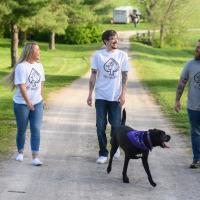 Cameron stands in between his friend Charlie, a white adult male with brown hair, and his fiance, Michelle, a white adult female with blonde hair. They are all wearing short sleeved specially designed ‘Team Cam’ shirts with a pair of jeans, while the dog walks ahead of them.