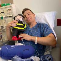Nick Corman, while in the hospital recovering from his kidney transplant, sits in bed with a teddy bear dressed like a fireman.