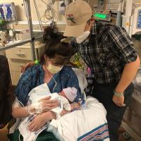 Sarah Custer, sitting, holds Isabel Custer, while flanked by Dave Custer, standing. They are all in a NICU.