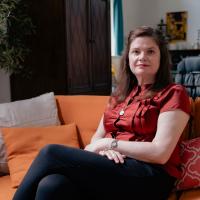 Nicola Allen, seated on a beige couch with three pillows, smiles softly at a camera.