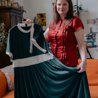 Nicola Allen, wearing a red shirt and a necklace, smiles toward camera while holding a black dress. She's standing in a room full of sewing materials.