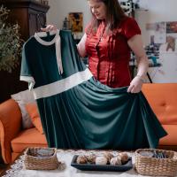 Nicola Allen, wearing a red shirt and standing, looks fondly at a black dress she's holding. She's standing in a room full of sewing materials.